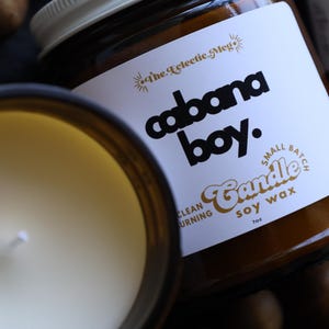 May include: A small batch soy wax candle in an amber glass jar with a white label. The label features the text 'The Eclectic Meg', 'cabana boy.', 'Small Batch', 'Candle', 'soy wax', and 'clean burning'. The candle has a white wick and 7oz weight.