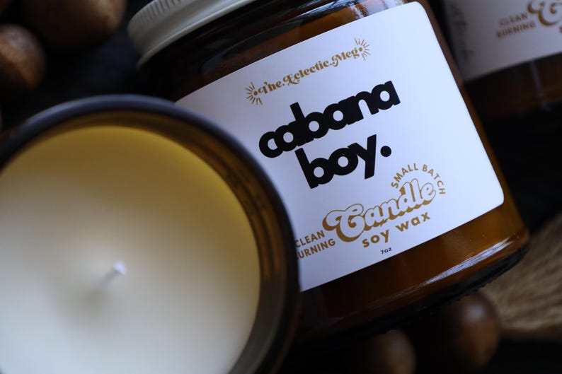 May include: A small batch soy wax candle in an amber glass jar with a white label. The label features the text 'The Eclectic Meg', 'cabana boy.', 'Small Batch', 'Candle', 'soy wax', and 'clean burning'. The candle has a white wick and 7oz weight.