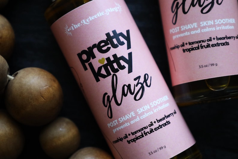 May include: A bottle of Pretty Kitty Glaze post shave skin soother with a pink label. The text reads: The Eclectic Meg, pretty kitty glaze, POST SHAVE SKIN SOOTHER, prevents and calms irritation, rosehip oil + tamanu oil + bearberry oil, tropical fruit extracts, 3.5 oz.