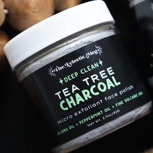 May include: Two jars of face polish with white lids and black labels. The labels read "Deep Clean Tea Tree Charcoal" in white and green text. The jars are 2.9oz/82g. The background is dark with brown orbs.