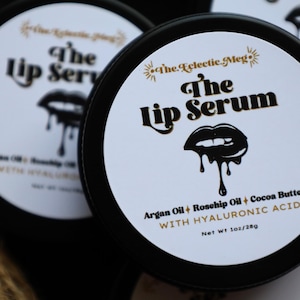 May include: A black jar with a white label that reads "The Lip Serum" in gold lettering. The label also includes the ingredients: argan oil, rosehip oil, cocoa butter, and hyaluronic acid. The jar is 1 oz / 28g.