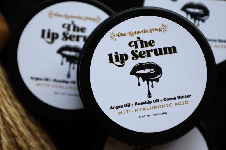 May include: A black jar with a white label that reads "The Lip Serum" in gold lettering. The label also includes the ingredients: argan oil, rosehip oil, cocoa butter, and hyaluronic acid. The jar is 1 oz / 28g.