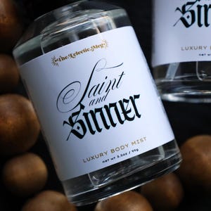 May include: Two clear glass bottles of "Saint and Sinner" luxury body mist with white labels and black gothic lettering. The bottles are surrounded by brown, round objects. The labels also include the text "net wt 3.5 oz / 99g".