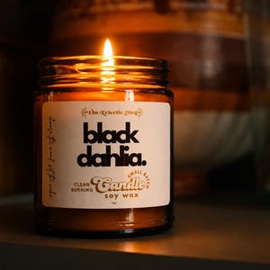 May include: A lit soy wax candle in a clear glass jar with a white label. The label features the text "The Eclectic Meg", "black dahlia.", "CLEAN BURNING", "Small Batch Candle", and "SOY WAX" in black and brown typography against a cream background.