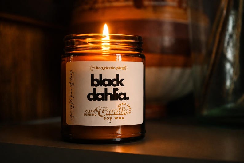 May include: A lit soy wax candle in a clear glass jar with a white label. The label features the text "The Eclectic Meg", "black dahlia.", "CLEAN BURNING", "Small Batch Candle", and "SOY WAX" in black and brown typography against a cream background.