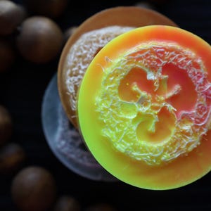 May include: Close-up of handmade soaps with a natural loofah center. The soaps are round, with vibrant colors of yellow, orange, and brown. The loofah adds texture and a natural element to the soaps. A dark background complements the colorful soaps.