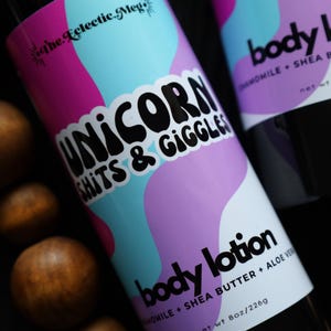 Könnte beinhalten: Zwei schwarze Körperlotion-Flaschen mit weißen Etiketten und farbenfrohen Designs. Auf einer Flasche steht "UNICORN SHITS & GIGGLES" und auf der anderen "body lotion". Beide Flaschen sind 226g.