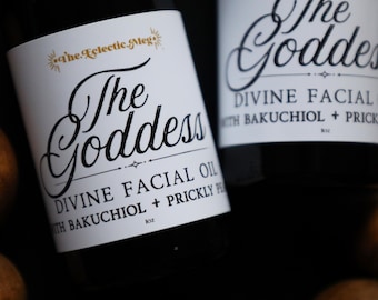 May include: Two dark glass bottles of facial oil with white labels. The labels feature the text The Eclectic Meg, The Goddess, Divine Facial Oil, with Bakuchiol + Prickly Pear, and 1oz. The bottles are surrounded by round, brown objects.