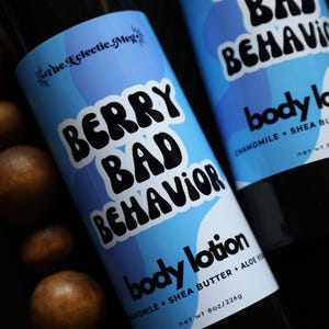 May include: Two bottles of body lotion with blue and white labels. The labels read "BERRY BAD BEHAVIOR" and "body lotion". The lotion contains chamomile, shea butter, and aloe vera. The bottles are surrounded by brown spheres.