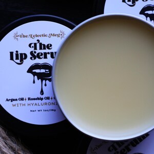 May include: A black and white lip balm container with the text "The Lip Serv" and "Argan Oil + Rosehip Oil + WITH HYALURONICS" on the label. The container is open and the lip balm is a light yellow color.