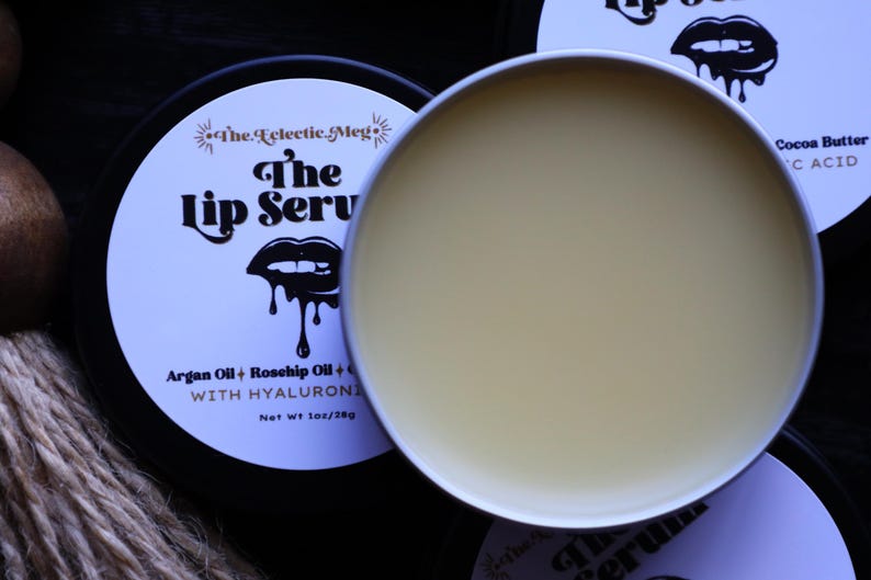 May include: A black and white lip balm container with the text "The Lip Serv" and "Argan Oil + Rosehip Oil + WITH HYALURONICS" on the label. The container is open and the lip balm is a light yellow color.