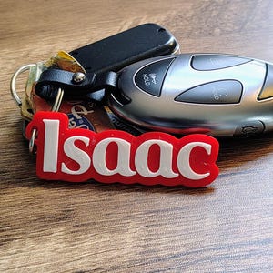 May include: A set of keys with a red and white "Isaac" keychain. The keys include a car key fob and a black key. The keychain is in a cursive font.