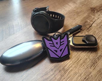 May include: A black smartwatch with a round face, a smooth dark gray oval case, a black key fob, and a keychain featuring a purple and black Decepticon logo emblem. These items are arranged together, showcasing a collection of personal accessories.