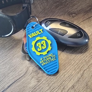 May include: A blue Vault 33 key tag with yellow accents, featuring the number 33 and the text "If Found Return to Vault-Tec." The tag is next to a car key fob and a black smartwatch.