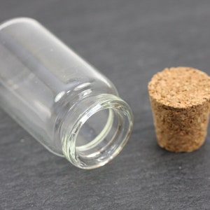 Könnte beinhalten: Eine klare Glasflasche mit einem Korkstopfen. Die Flasche ist leer und hat eine runde Form.