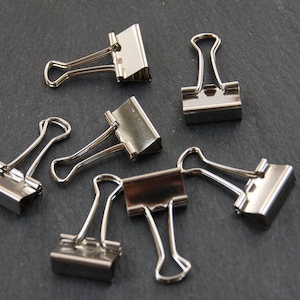 Pode incluir: Seis clipes de pasta de arquivo prateados sobre uma superfície cinza escura. Os clipes estão todos fechados e dispostos em um padrão aleatório.