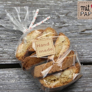 Può includere: Un sacchetto di plastica trasparente pieno di diversi biscotti biscotti. Il sacchetto è legato con un nastro bianco con pois rossi. Un'etichetta di carta marrone con il testo "mit ♥ & PAPIER" si trova nell'angolo in alto a destra dell'immagine. Un'etichetta di carta marrone più piccola con il testo "Hand made" è attaccata al sacchetto.