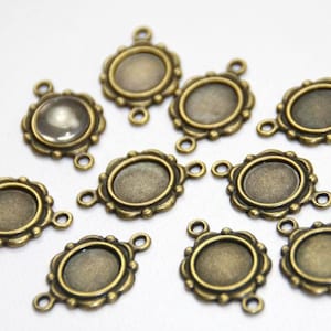Pode incluir: Uma variedade de bases de pingente ovaladas em tom bronze antigo. Cada base tem uma borda decorativa ondulada e duas pequenas argolas para fixação. Uma base tem um cabochão de vidro transparente.