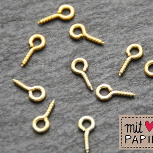 May include: Gold-toned screw-in eyelet findings for jewelry making. The eyelets have a small loop at the top and a screw-in post at the bottom.