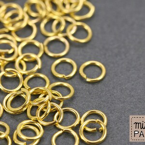 Könnte beinhalten: Eine Ansammlung kleiner, kreisförmiger goldfarbener Metall-Biegeringe, die über den Rahmen verstreut sind. Jeder Ring weist einen einzelnen offenen Schlitz auf, der die Verbindung bei der Schmuckherstellung ermöglicht. Ein Logo zeigt mit & PAPIER.