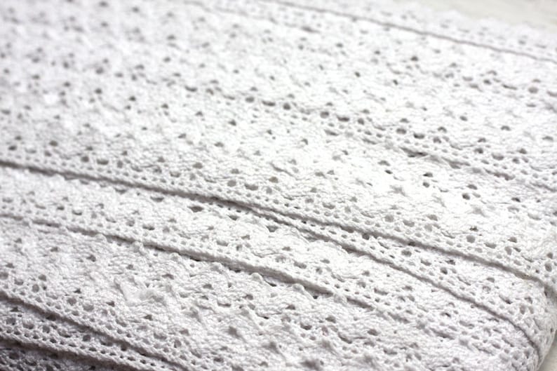 May include: White cotton lace trim with a delicate floral pattern. The lace is woven in a repeating pattern of small flowers and leaves.