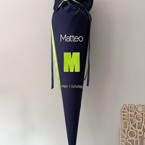 Könnte beinhalten: Eine dunkelblaue Schultüte mit dem Namen "Matteo" in Weiß, einem großen grünen "M" und dem Text "Mein 1. Schultag". Die Tüte ist oben mit einem Kordelzug gebunden und hat grüne Bänder. Ein Holzalphabet befindet sich im Hintergrund.