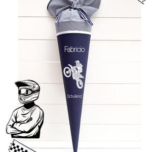 Könnte beinhalten: Eine dunkelblaue, kegelförmige Schultüte mit einem oben mit Band gebundenen marineblau-weiß karierten Stoffabschluss. Die Tüte zeigt den Namen Fabricio, eine weiße Grafik einer Person auf einem Motocross-Motorrad und den Text Schulkind. Inklusive Motocross-Helm.