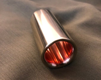 Puede incluir: Un slide de guitarra plateado con un interior de cobre. El slide es de forma cilíndrica y está hecho de metal.
