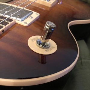 May include: Close-up of a brown and black electric guitar with a cream-colored pickguard. The guitar has a bullet-shaped output jack with a cream-colored ring around it. The text "PUSH" is printed on the ring.