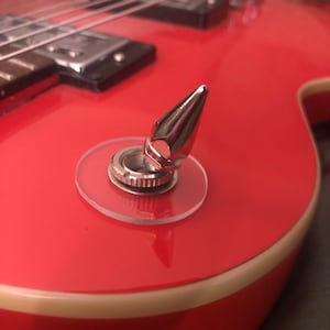 Puede incluir: Primer plano de una guitarra eléctrica roja con una toma de salida cromada y una cubierta de plástico transparente. La guitarra tiene una encuadernación blanca alrededor del borde.