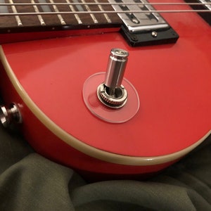 Może przedstawiać: Zbliżenie czerwonej gitary elektrycznej z chromowanym przełącznikiem i przezroczystą osłoną z tworzywa sztucznego. Gitara ma kremową obwódkę wokół krawędzi.