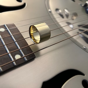 May include: A gold-colored metal guitar slide rests on the strings of a silver resonator guitar. The fretboard is dark brown with white fret markers. The slide is a cylindrical shape.