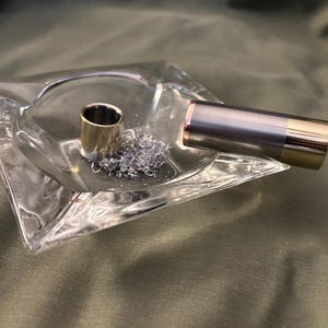 May include: A clear glass ashtray with a silver metal cylinder and a gold metal cylinder inside. The ashtray is on a green fabric surface.