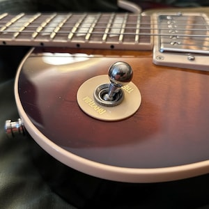 Op de afbeelding: Close-up van een elektrische gitaar met een sunburst-afwerking. De gitaar heeft een chromen tuimelschakelaar met de tekst "Rhythm" en "Treble" op een crèmekleurige plaat. De snaren en toets zijn zichtbaar, samen met een chromen pickup.