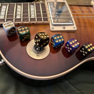 May include: A collection of six dice with gold-colored dots, resting on a brown and tan electric guitar. The dice are in various colors, including black, blue, green, and purple. The guitar's strings and pickups are also visible.