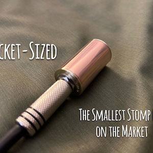 Pode incluir: Um pequeno pedal de efeitos de cor prata e ouro rosa com o texto "POCKET-SIZED" e "THE SMALLEST STOMP BOX ON THE MARKET" impresso no fundo.