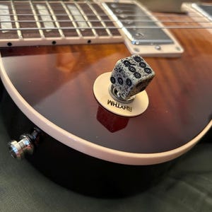 May include: Close-up of an electric guitar with a brown and black finish. A dice-shaped knob with black circle designs sits on a cream-colored base labeled "RHYTHM". The guitar's strings and pickups are visible, with a light-colored edge.