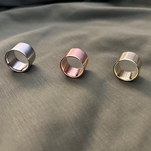 May include: Three metal rings in different finishes: silver, rose gold, and gold. The rings are thick and have a cylindrical shape.