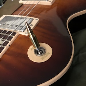 Pode incluir: Um close-up de uma guitarra elétrica marrom e dourada com um interruptor de alavanca em forma de bala. O interruptor está etiquetado como "Gibson".