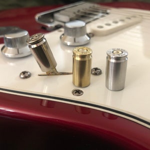 Puede incluir: Tres casquillos de bala se utilizan como perillas en una guitarra eléctrica blanca. Los casquillos son de latón, plata y níquel. La guitarra es roja.