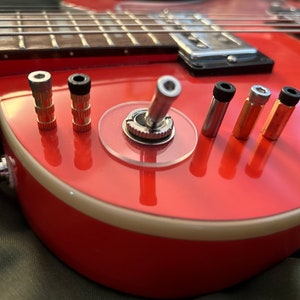 Peut inclure: Gros plan sur une guitare électrique rouge avec un interrupteur à bascule chromé et plusieurs prises de sortie dorées et argentées.