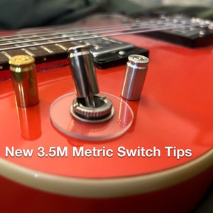 以下が含まれることがあります： 赤い電気ギターのアップで、スイッチの上に透明なプラスチックカバーがかけられています。スイッチには銀色のノブと銀色の先端が付いています。ギターのボディには銀色の金属製の先端が2つ付いています。ギターの下には「New 3.5M Metric Switch Tips」という文字が見えます。