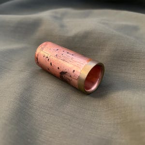May include: A copper ring with a brass band. The ring has a cylindrical shape with a reddish-brown hue and dark spots. The brass band is a gold color. The ring is on a green fabric.