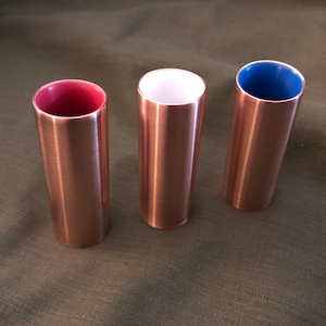 May include: Three copper shot glasses with different colored interiors. The glasses are lined with red, white, and blue.