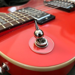 Può includere: Primo piano di una chitarra elettrica rossa con un interruttore a levetta cromato e un bordo bianco.