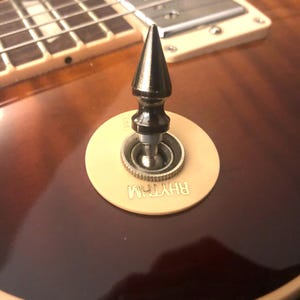May include: A close-up of a guitar's rhythm switch with a black, pointed knob. The switch is labeled "RHYTHM" in gold lettering.