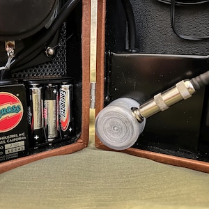 Puede incluir: Un amplificador de guitarra Pignose marrón y negro con un altavoz, una entrada jack y un compartimento para baterías con cuatro baterías AA. El amplificador está etiquetado como "Pignose Industries, Inc. Los Ángeles, California. Modelo n.º 7-100. N.º de serie 48841."