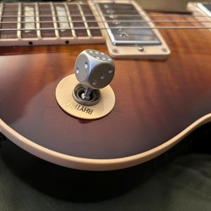 Può includere: Primo piano di una chitarra elettrica con finitura marrone e ambra. Un interruttore argentato a forma di dado è montato su una placca color crema con la scritta "RHYTHM". Sono visibili le corde e la tastiera della chitarra.