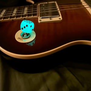 May include: A close-up of an electric guitar with a brown finish. A glowing blue dice-shaped switch tip is installed on the guitar. The guitar's fretboard and pickups are visible.