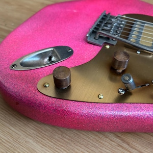 Op de afbeelding: Close-up van een roze elektrische gitaar met een glitterafwerking. De gitaar heeft een messingkleurige slagplaat, metalen knoppen en een zilveren brug. De body heeft een afgeronde vorm en de snaren zijn zichtbaar.